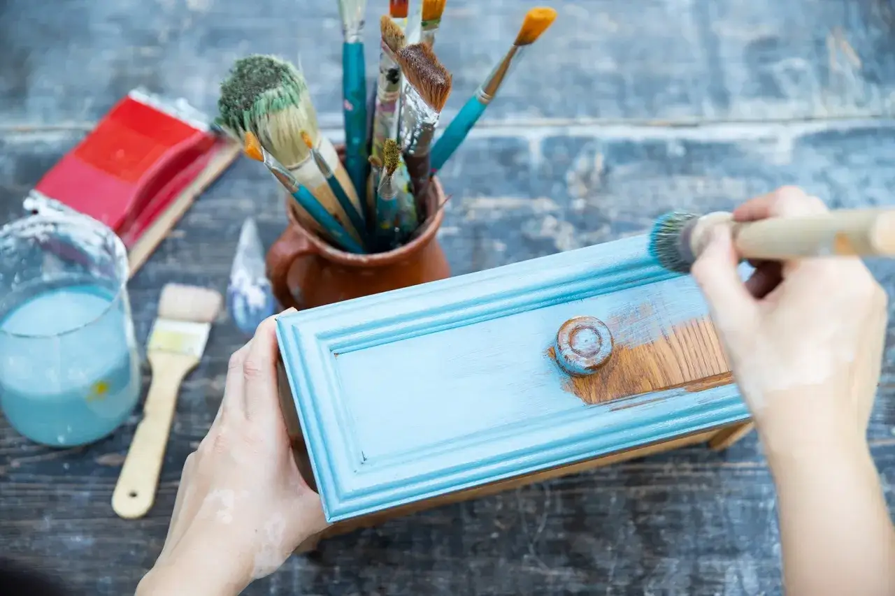 Ręce malują niebieską szufladę, odnawiając meble. Pędzle i farby czekają na dalsze szlifowanie mebli.