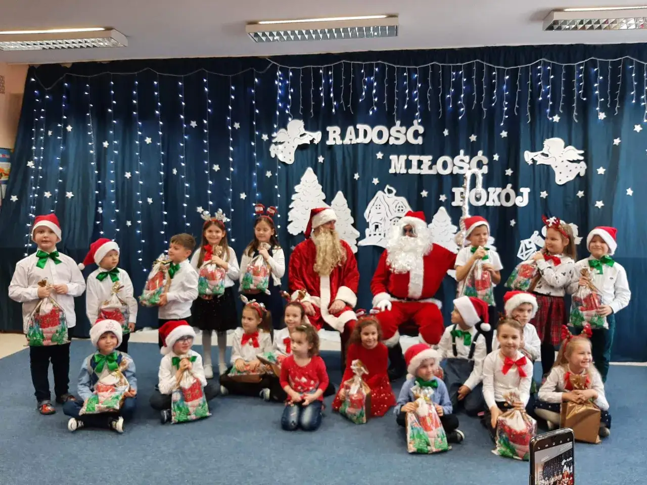 Wesołe **święta w przedszkolu**! Dzieci w strojach mikołajkowych z prezentami pozują do zdjęcia z Mikołajami.
