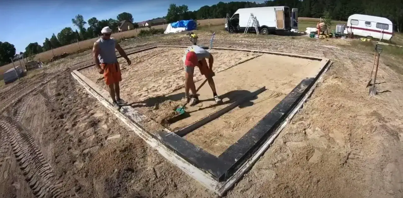 Budowa fundament&oacute;w pod dom 70m2. Dw&oacute;ch mężczyzn przygotowuje teren, jeden kopie łopatą, drugi nadzoruje. W tle przyczepa kempingowa i samoch&oacute;d dostawczy.