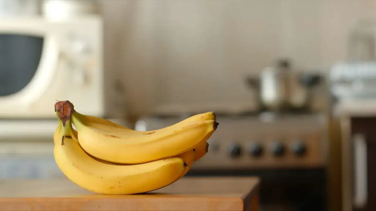 Pęczek ż&oacute;łtych banan&oacute;w na drewnianym blacie. W tle kuchenka i mikrofal&oacute;wka. Dowiedz się, jak przechowywać banany, by dłużej były świeże.