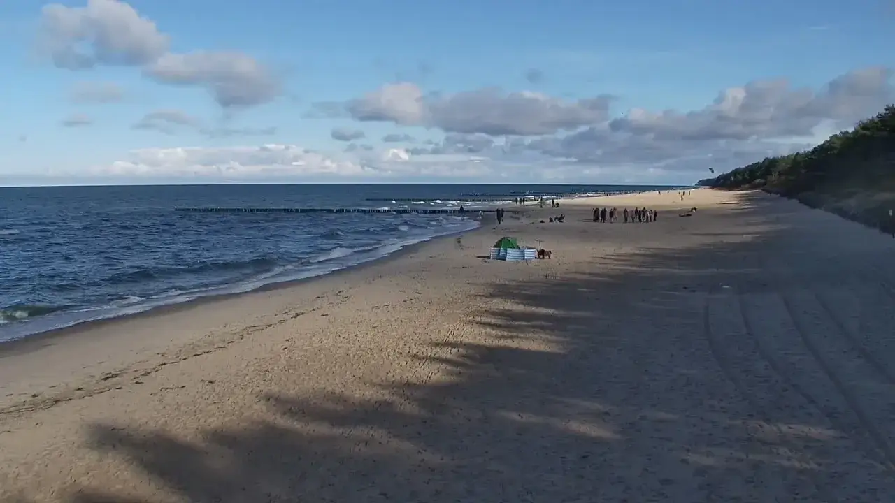 Pobierowo atrakcje: szeroka plaża, morze, falochrony i spacerujący ludzie. W tle las.