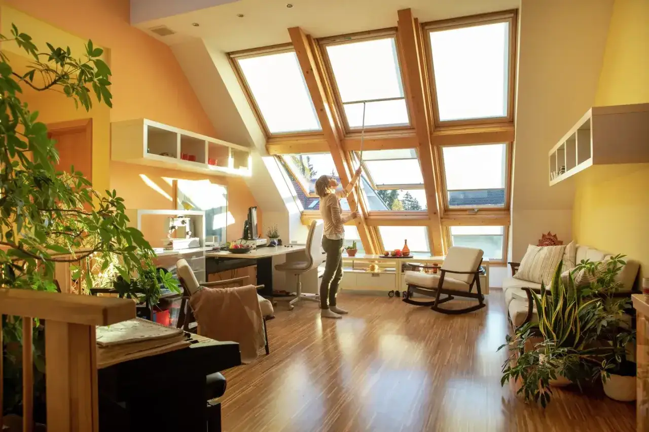 Kobieta otwiera okna dachowe, zapewniając doświetlenie pomieszczeń światłem naturalnym. Wnętrze jest jasne i przytulne, z roślinami i meblami.