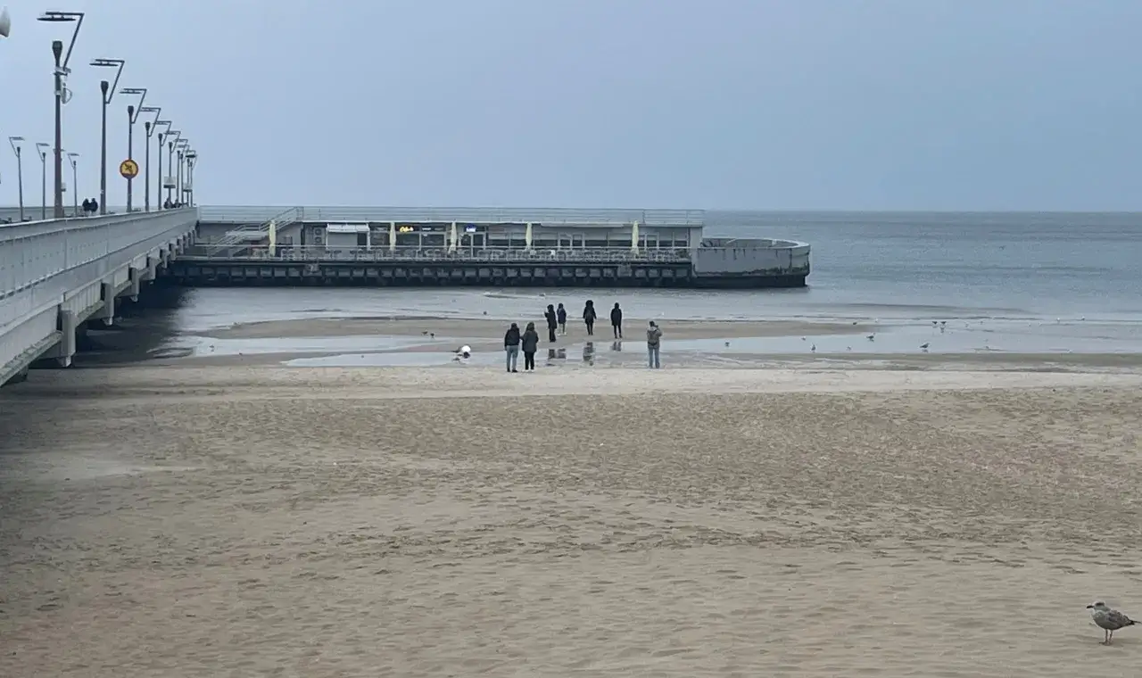 Spacerem po plaży w Kołobrzegu, zastanawiając się, czy molo w Kołobrzegu jest płatne. W oddali widać budynek i morze.