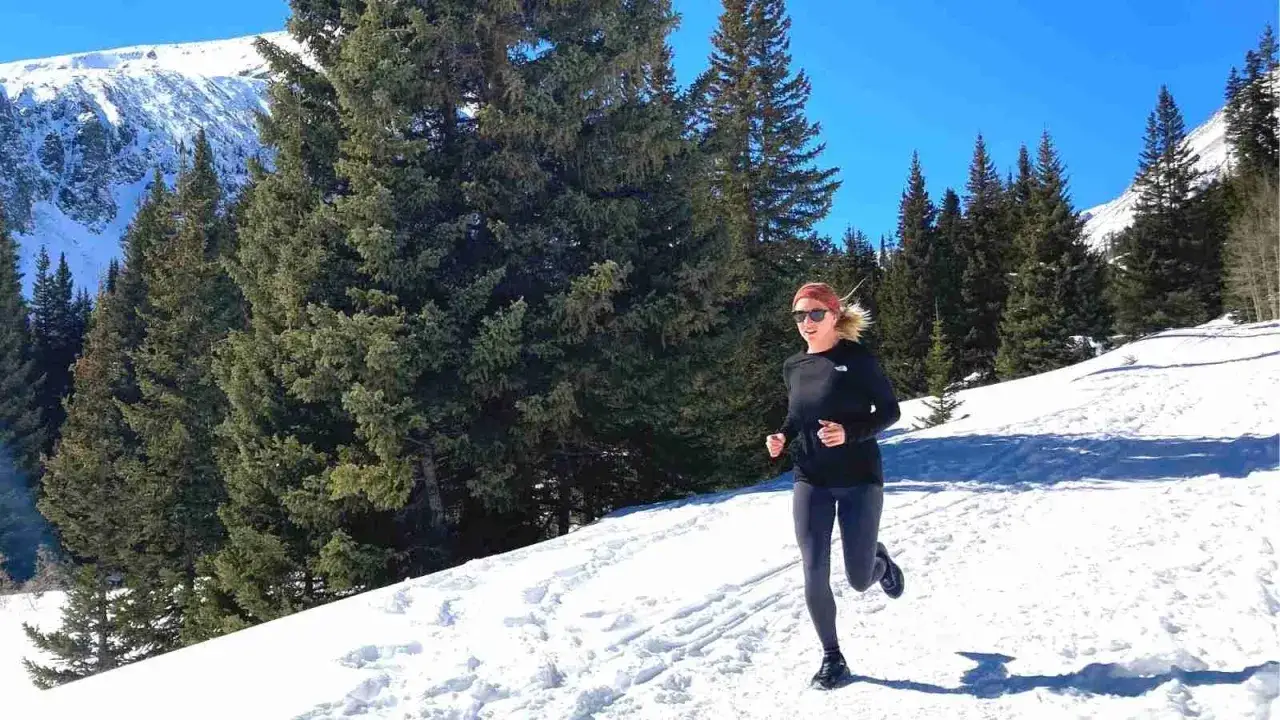 Kobieta biegnie po ośnieżonym szlaku wśr&oacute;d drzew. Pokazuje, jak biegac w zime, ciesząc się zimową scenerią g&oacute;r.