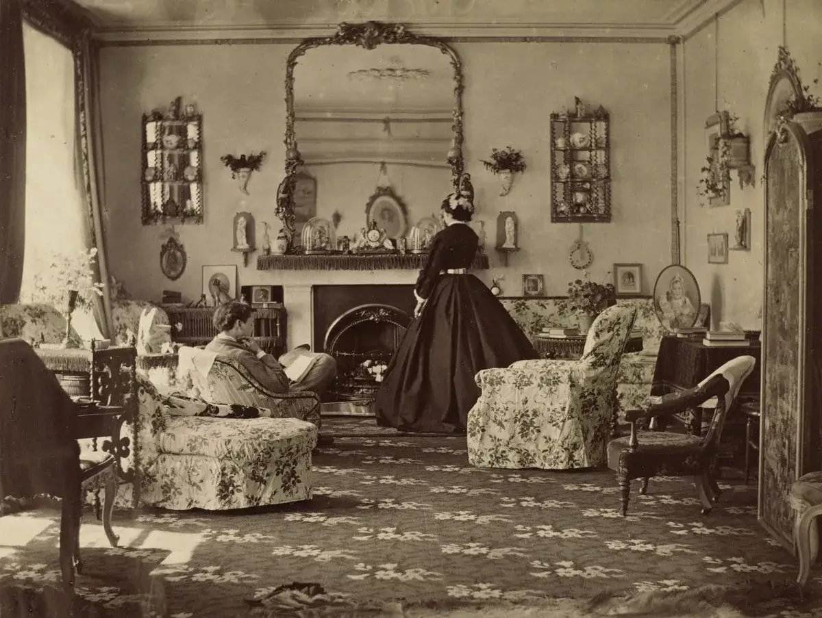 Elegancka kobieta w czarnej sukni stoi w salonie na plebanii w Haworth. Mężczyzna czyta książkę, siedząc w fotelu.