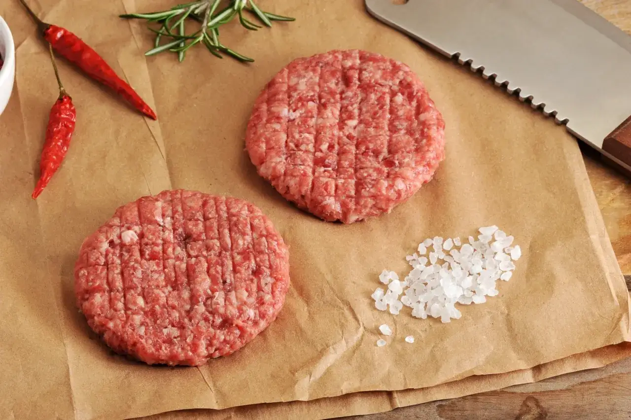 Dwa surowe kotlety na burgery, przyprawy i nóż. Idealne składniki, by dowiedzieć się, jak zrobić burgery jak zrobic mięso.