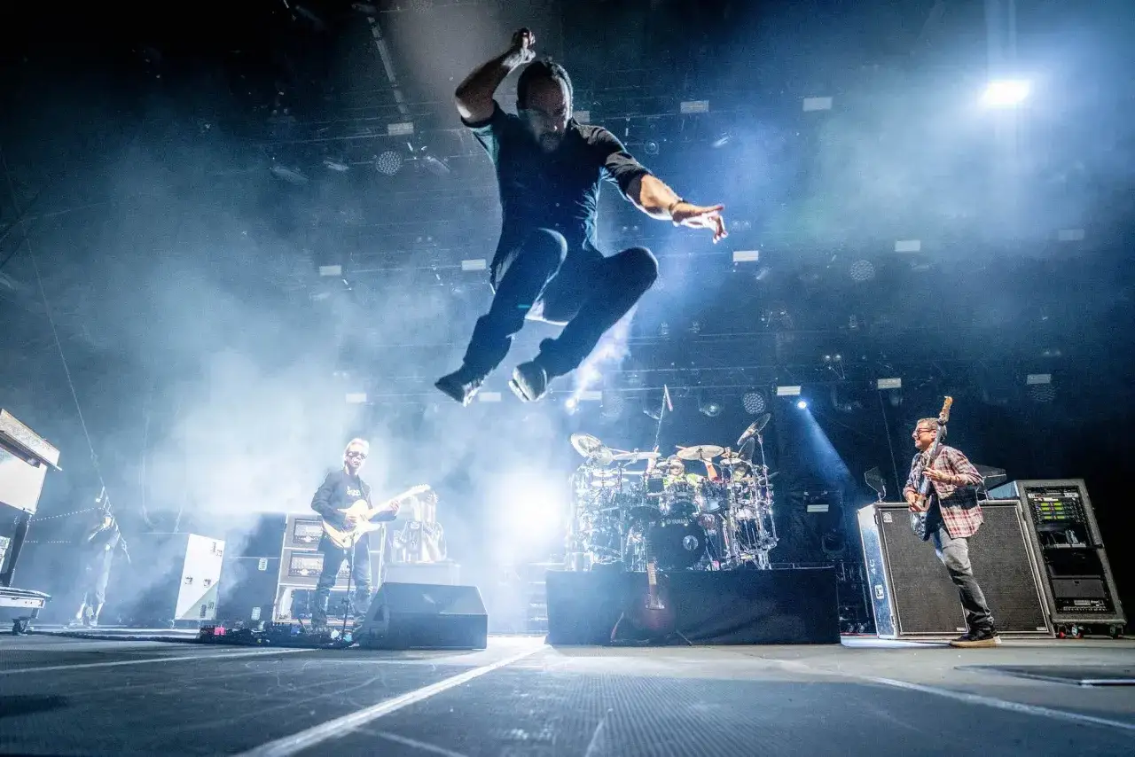 Energiczna fotografia koncertowa: wokalista w powietrzu, gitarzyści i perkusista na scenie.