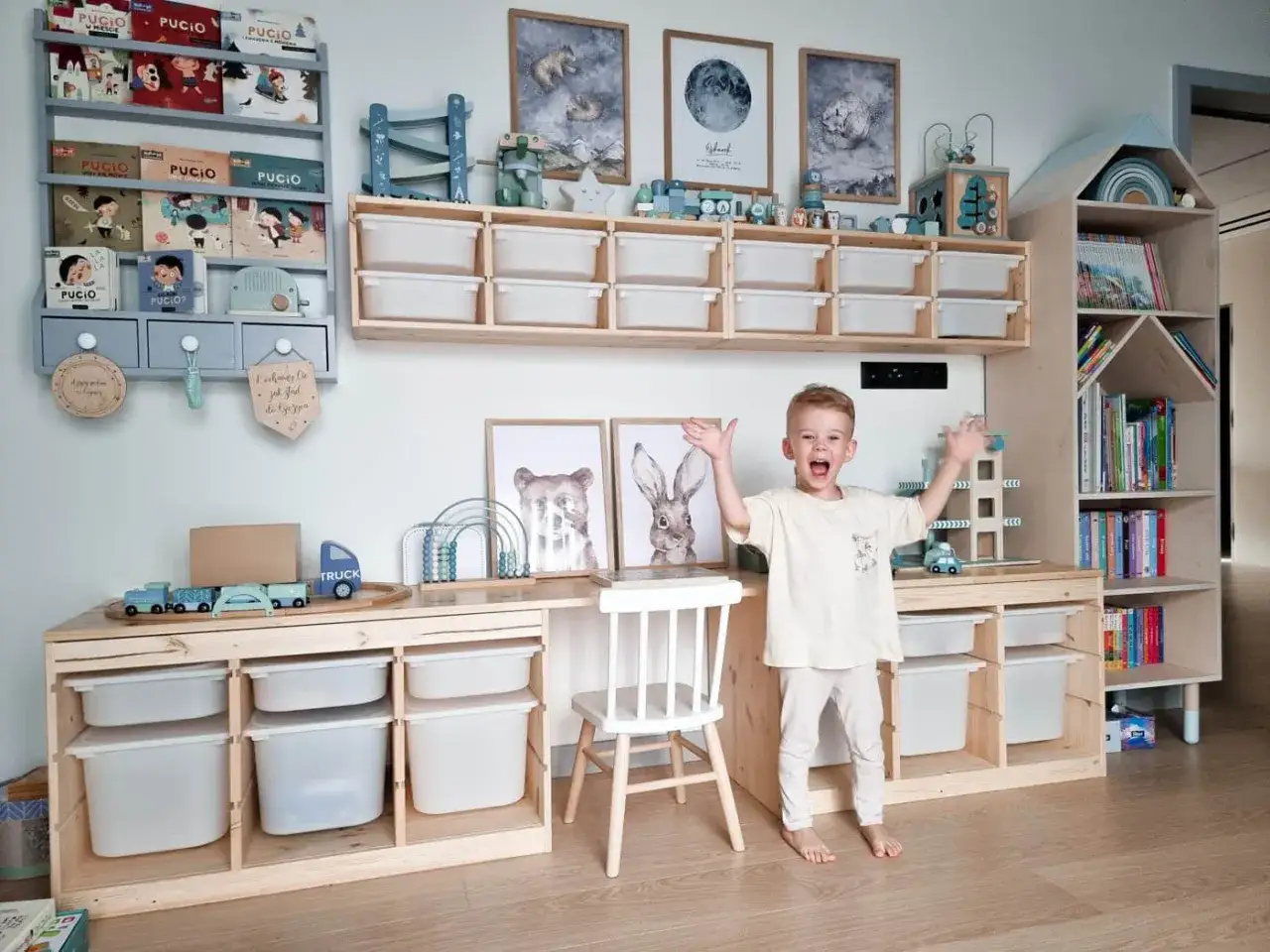 Radosny chłopiec 4 lata w swoim pokoju pełnym zabawek i książek. Drewniane meble, regały z organizerami i dekoracje tworzą przytulną przestrzeń.