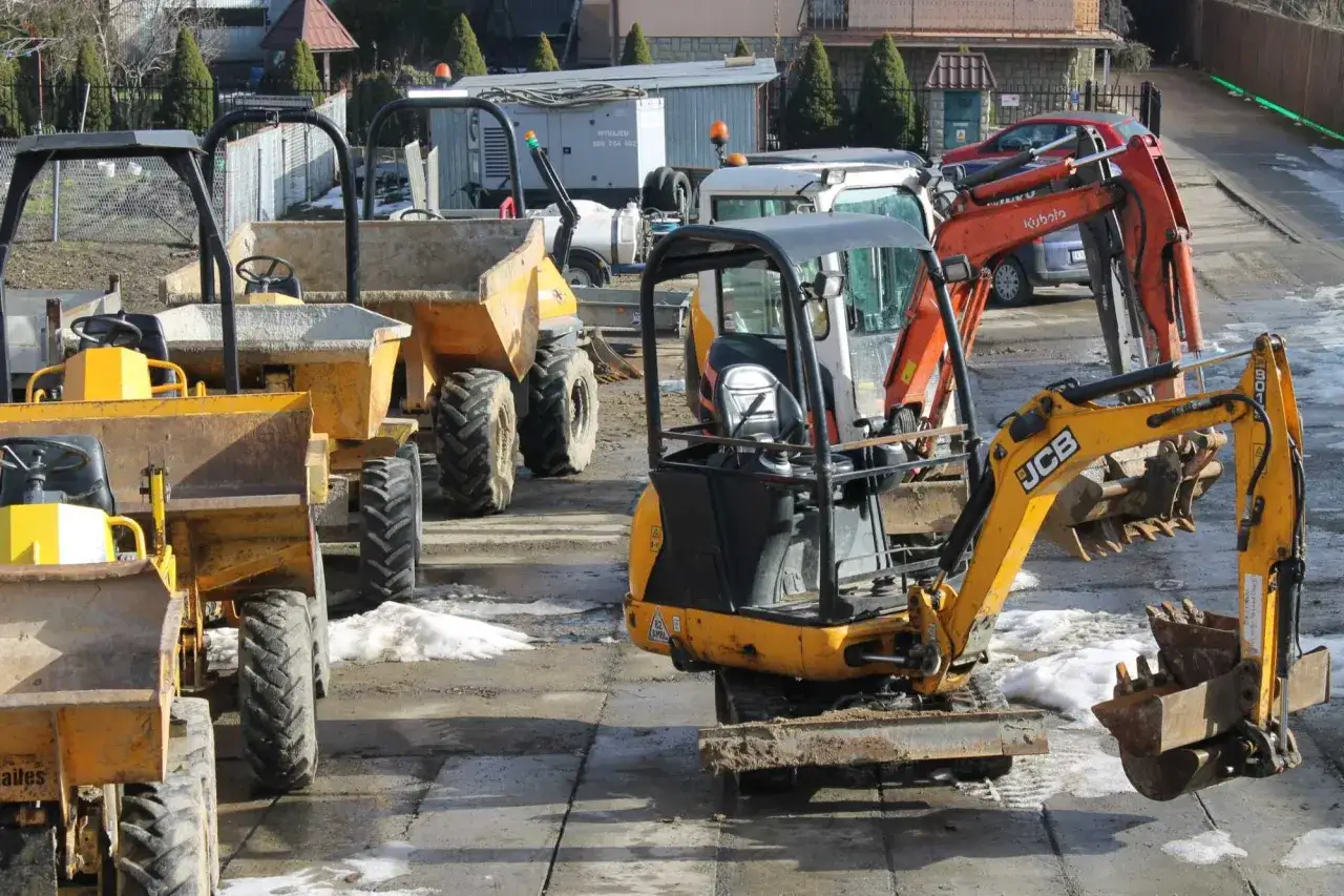 Kilka żółtych i pomarańczowych koparek oraz wywrotek stoi na placu budowy. Zastanawiasz się, ile kosztuje wynajem koparki?