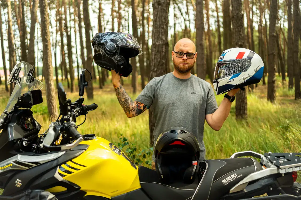 Mężczyzna z tatuażami por&oacute;wnuje kask szczękowy i integralny, stojąc obok ż&oacute;łtego motocykla Suzuki w lesie.