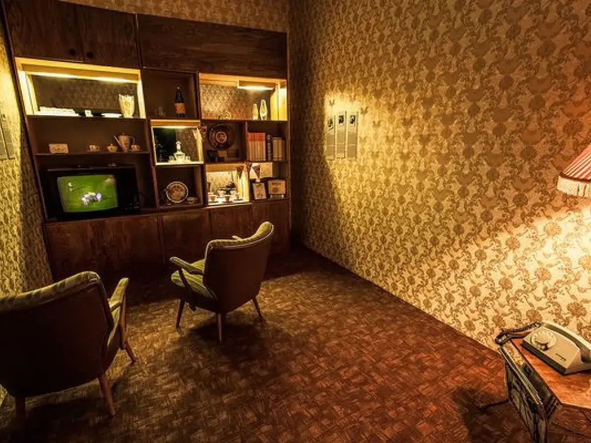 Wnętrze w stylu pok&oacute;j lata 80. z meblościanką, telewizorem, dwoma fotelami i telefonem na stoliku.