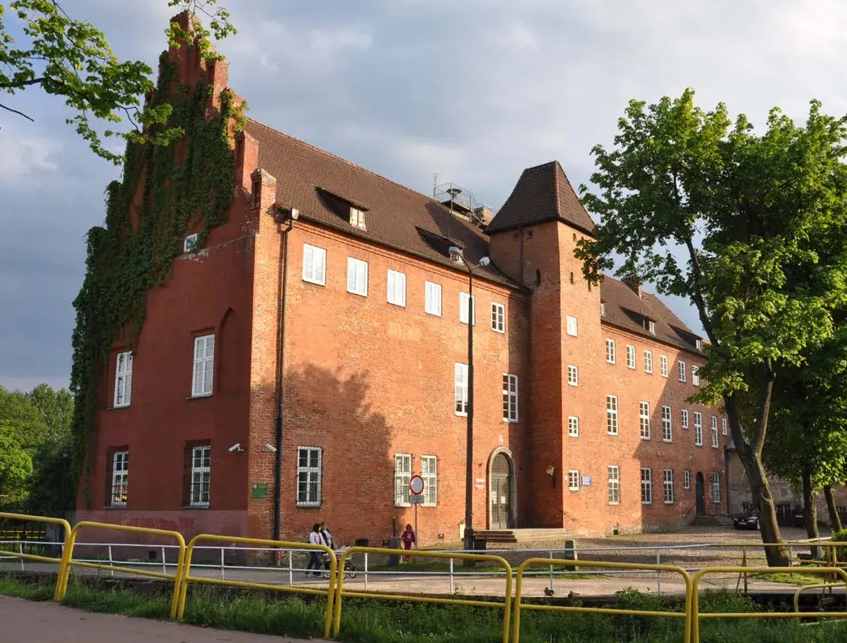 Zamek krzyżacki w Lęborku, ceglana budowla z wieżą i zielenią pnącą się po ścianach, otoczona żółtym ogrodzeniem.