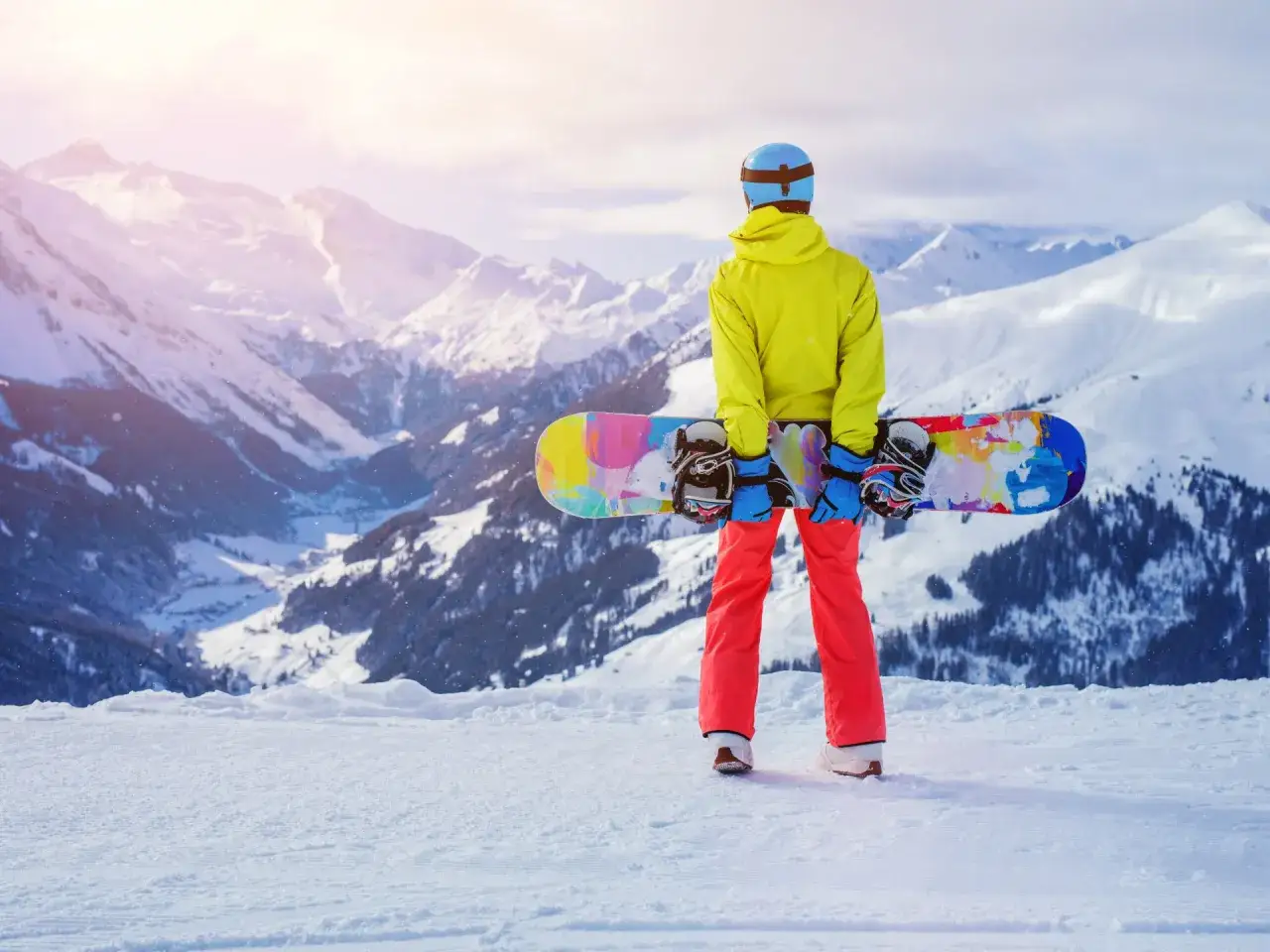 Snowboardzista w jaskrawej kurtce i czerwonych spodniach trzyma kolorowego snowboarda, gotowy do zjazdu. Widok na ośnieżone góry. Co to jest skipass? To klucz do tej przygody!