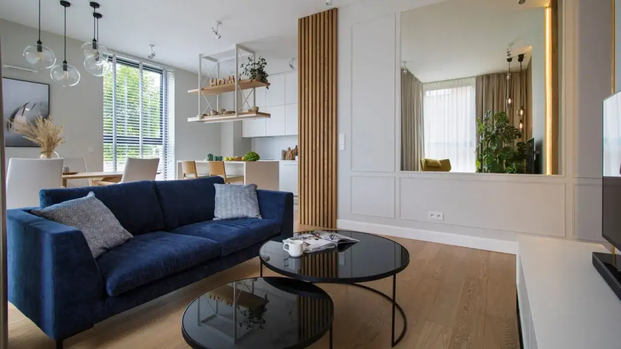 Nowoczesny salon z granatową kanapą i czarnymi stolikami. Duże lustro na ścianie odbija rośliny i zasłony, co oznacza zbicie lustra w kontekście przesądów.