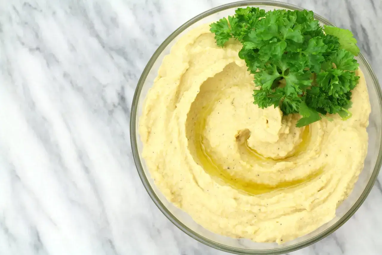 Puszysty hummus z ciecierzycy w szklanej misce, polany oliwą i ozdobiony gałązką natki pietruszki, na marmurowym tle.