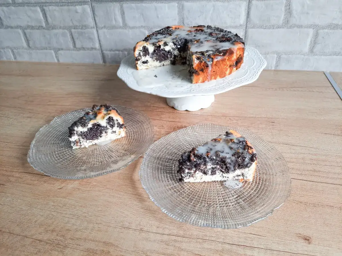 Pyszny sernik z makiem, polany lukrem, na białej paterze i dwóch talerzykach.