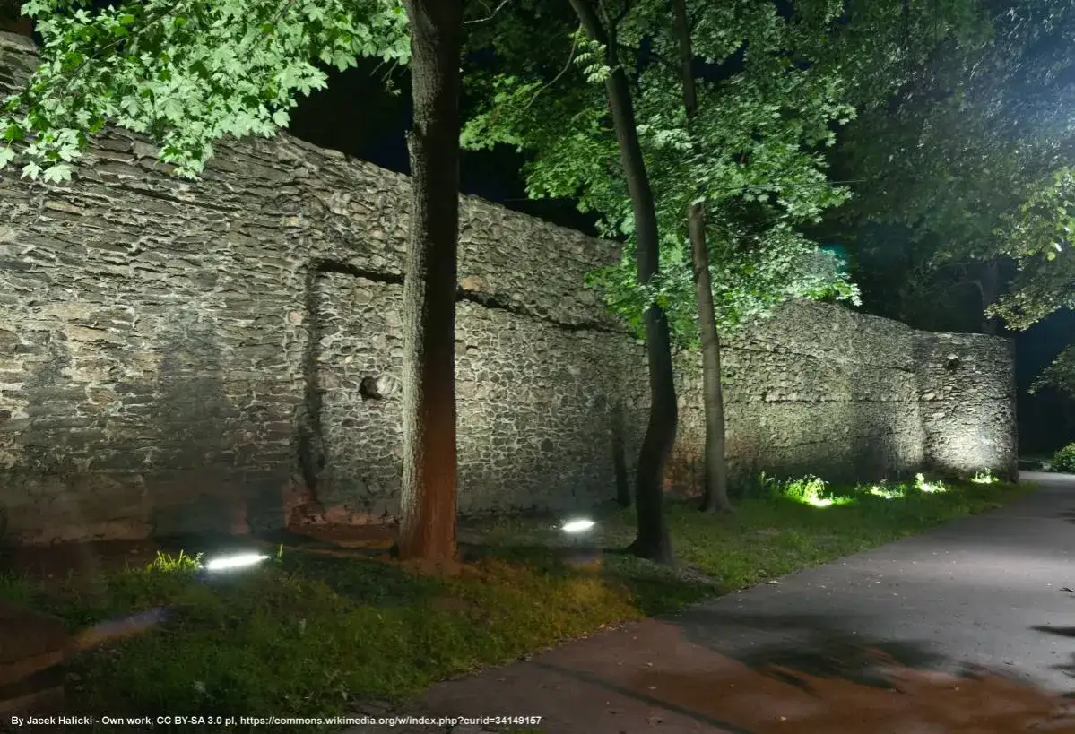 Nocne paczk&oacute;w atrakcje: kamienne mury zamku oświetlone punktowo, drzewa rzucają cienie.
