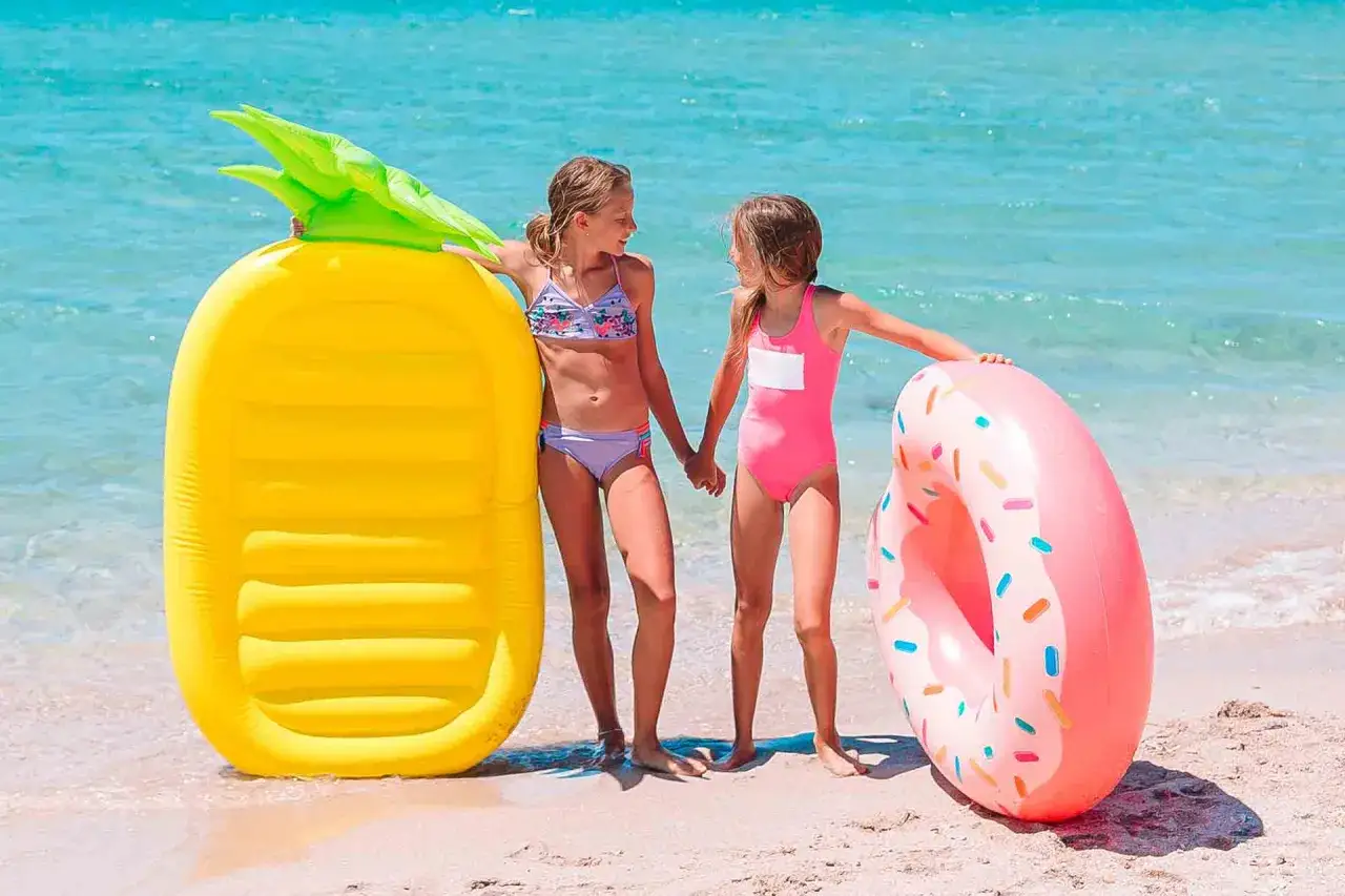 Dwie dziewczynki trzymają się za ręce na plaży obok żółtego materaca do pływania w kształcie ananasa i różowej dmuchanej opony w kształcie pączka.