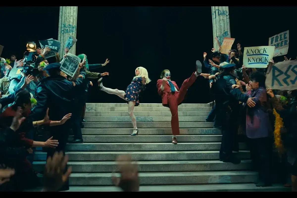 Joker i Harley Quinn tańczą na schodach wśród tłumu z transparentami. Recenzje Joker: Folie à Deux zapowiadają szaleństwo.