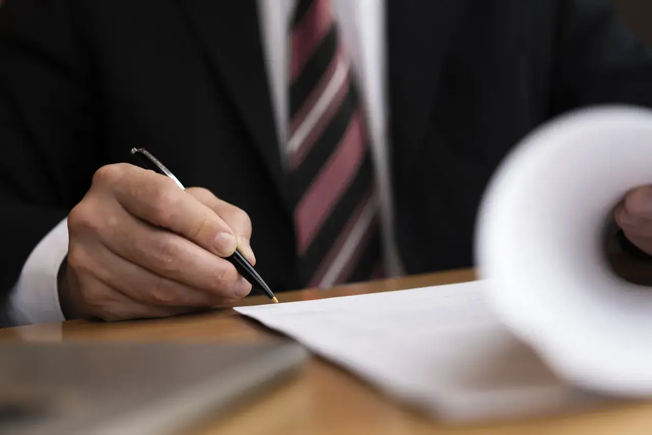 Mężczyzna w garniturze podpisuje umowę przedwstępną sprzedaży domu. Długopis i dokumenty na biurku.
