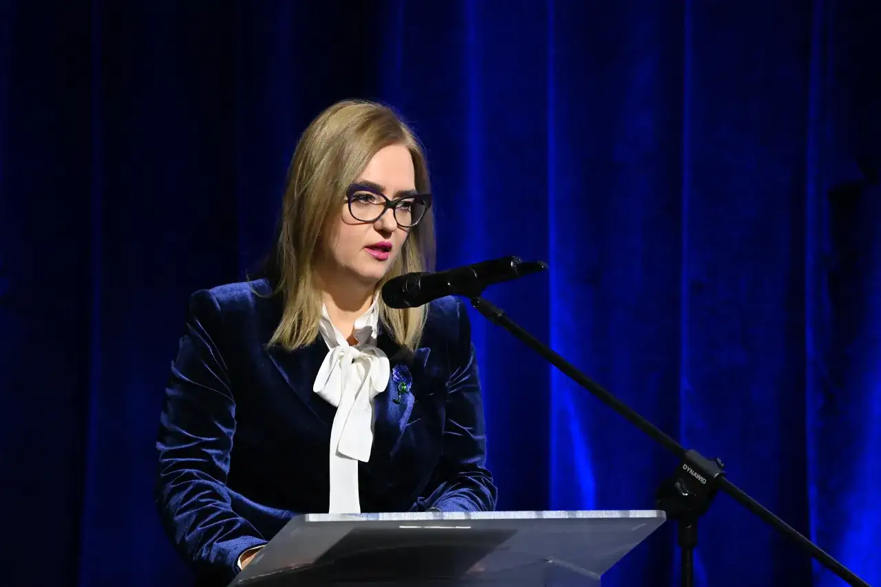 Magda Adamowicz, w eleganckim granatowym żakiecie i białej bluzce, przemawia przy mównicy. Jej wiek nie jest widoczny, ale pewność siebie tak.