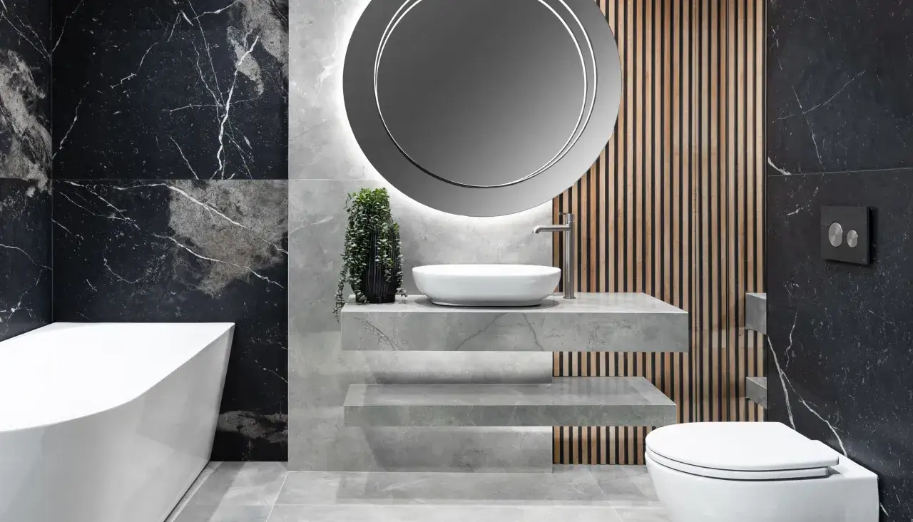 Nowoczesny blat pod umywalkę z płytek, z wiszącą rośliną i okrągłym lustrem, w łazience z wanną i toaletą.