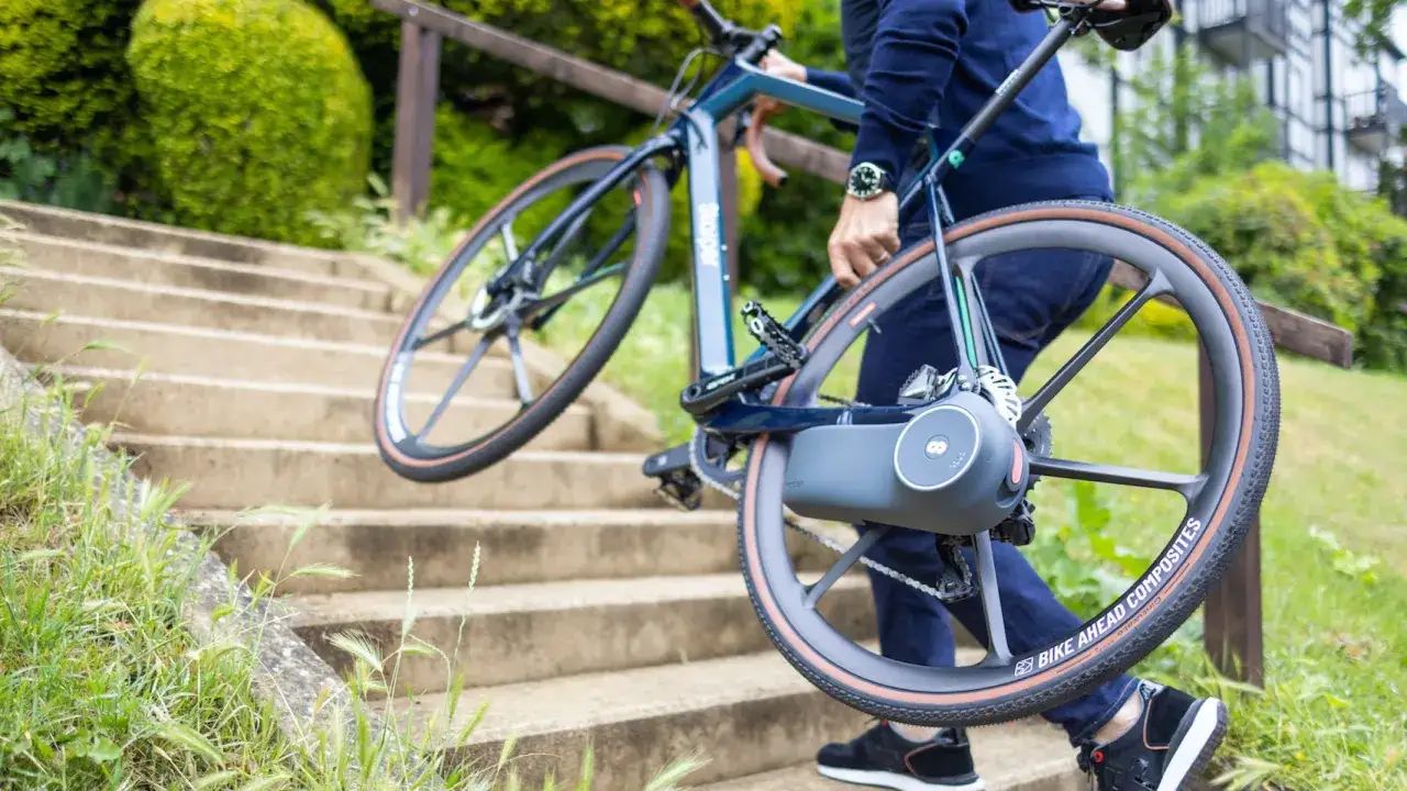 Osoba wnosi rower po schodach, prezentując nowoczesne gadżety do roweru, w tym innowacyjny napęd w tylnym kole.