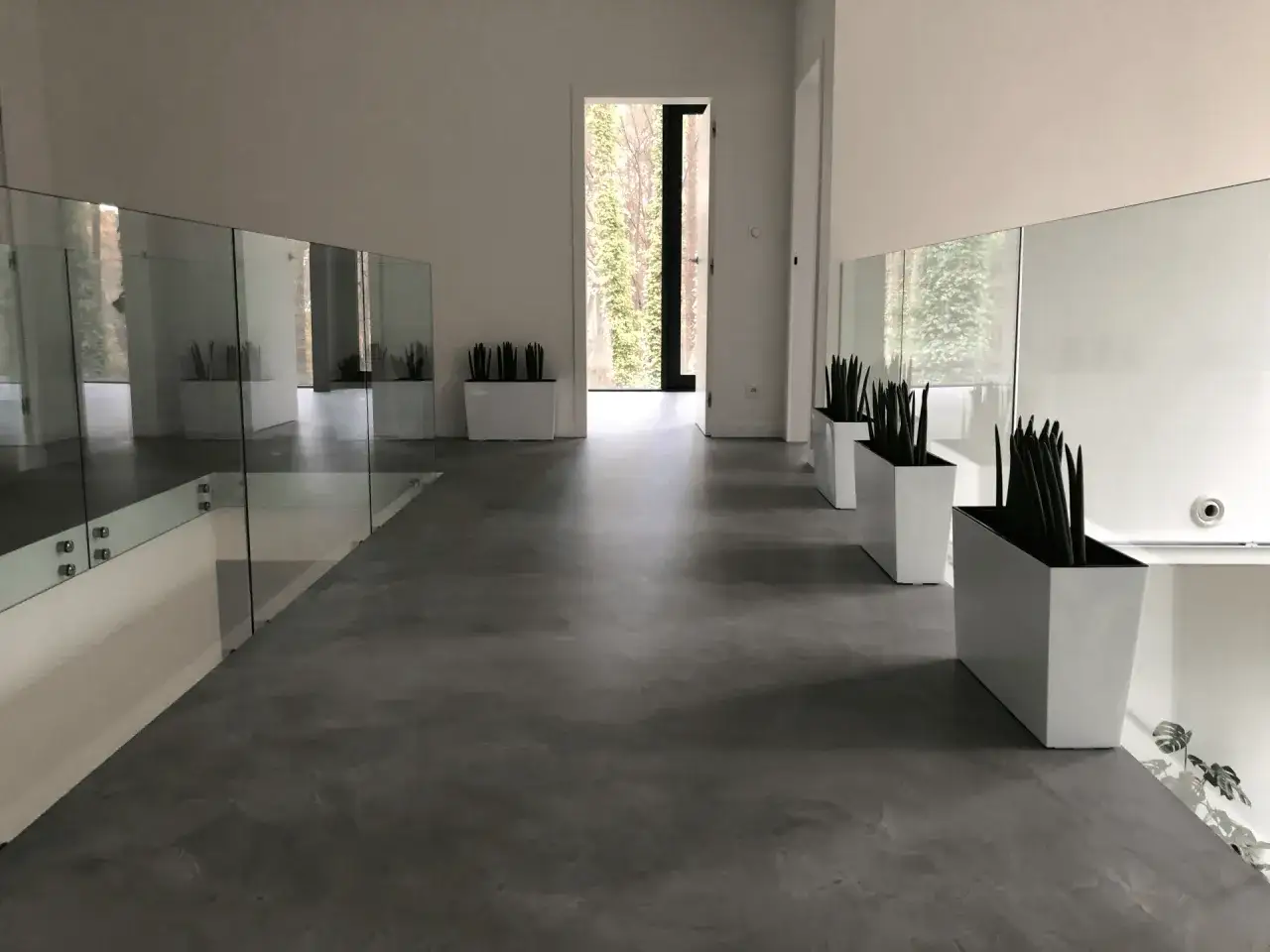 Nowoczesne wnętrze z gładkimi posadzkami z mikrocementu, szklanymi balustradami i białymi donicami z roślinami.