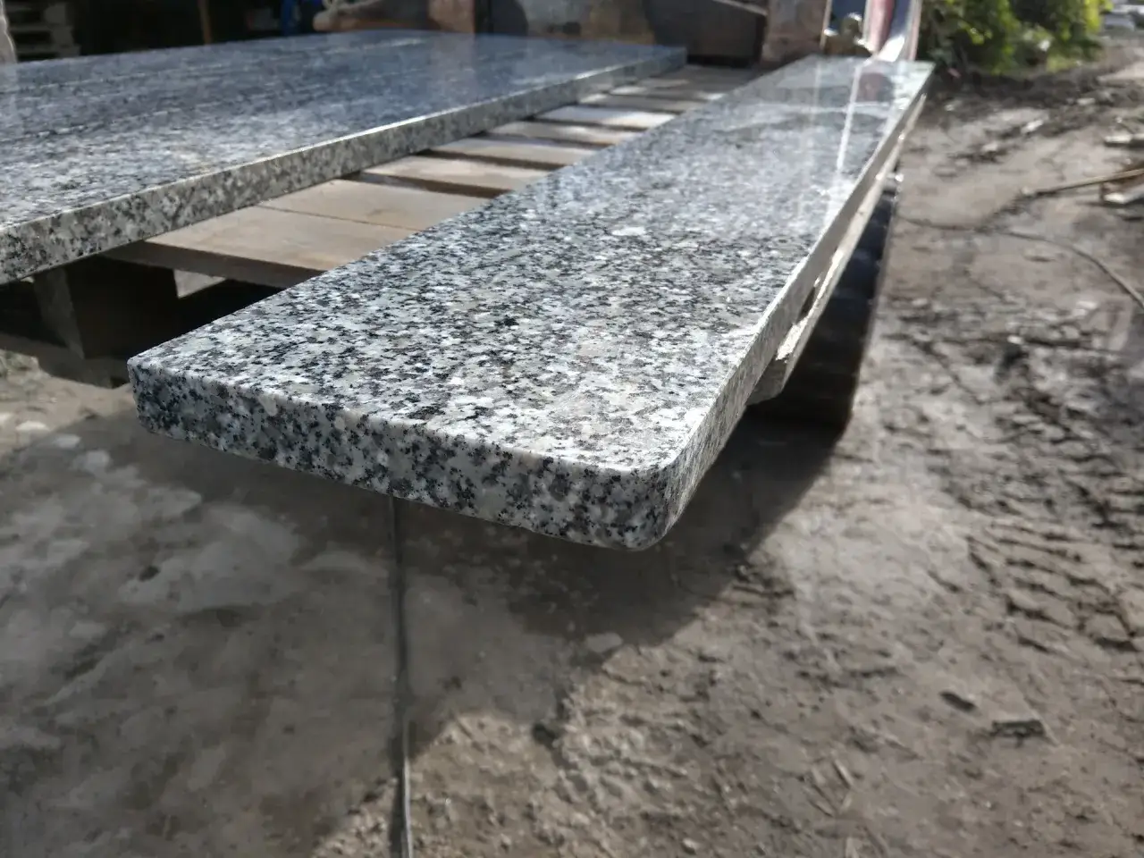 Srebrzysty granit o drobnej, plamistej fakturze, przypominający szary piryt, leży na drewnianej palecie.