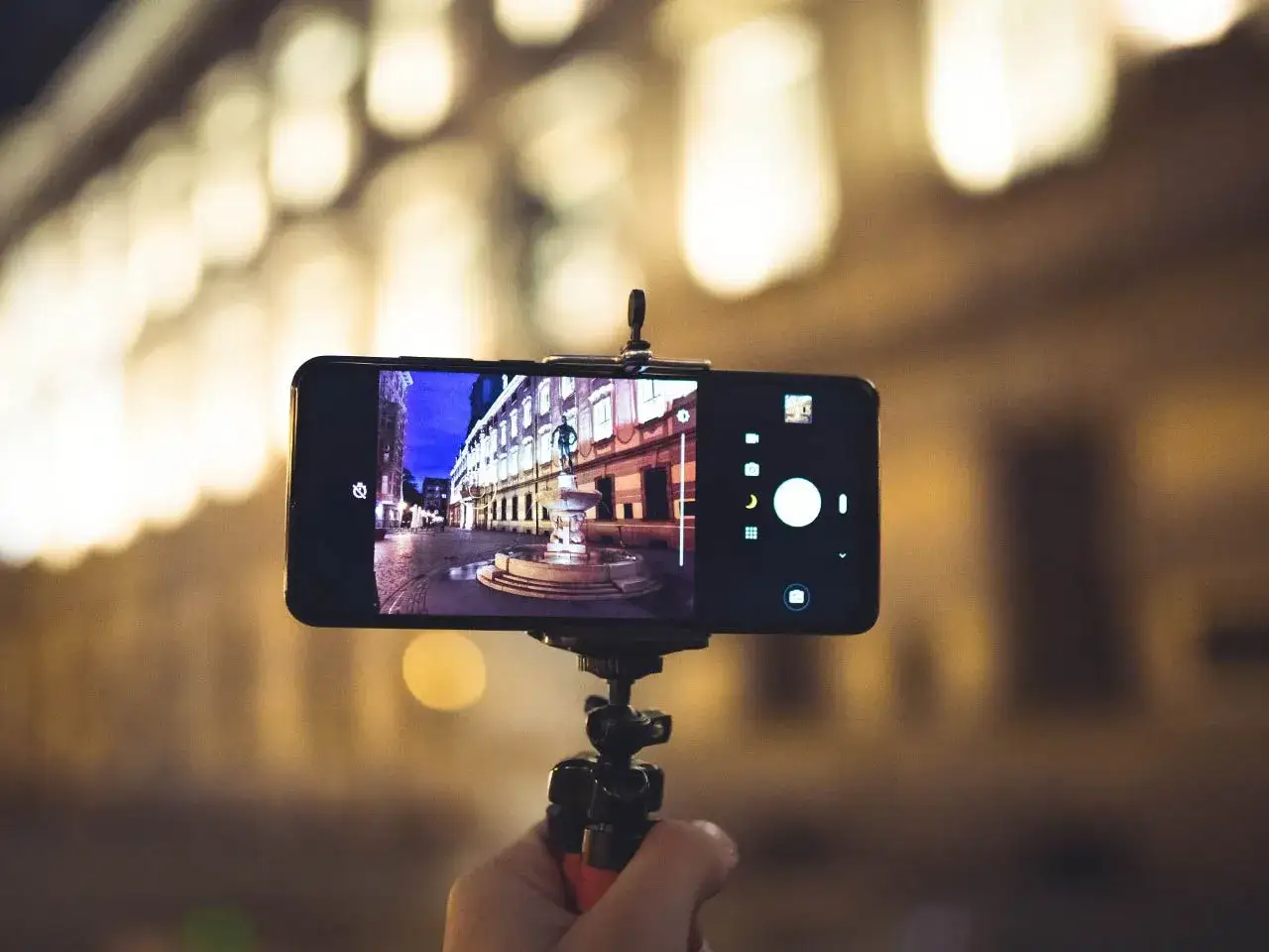 Telefon na statywie rejestruje nocną scenę z fontanną. Dowiedz się, jak ustawić długi czas naświetlania w telefonie, by uzyskać takie efekty.