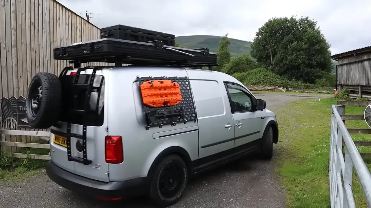 Srebrny caddy kamper z zamontowanym na dachu boxem i kołem zapasowym z tyłu. Na bocznych drzwiach pomarańczowe wyciągarki.