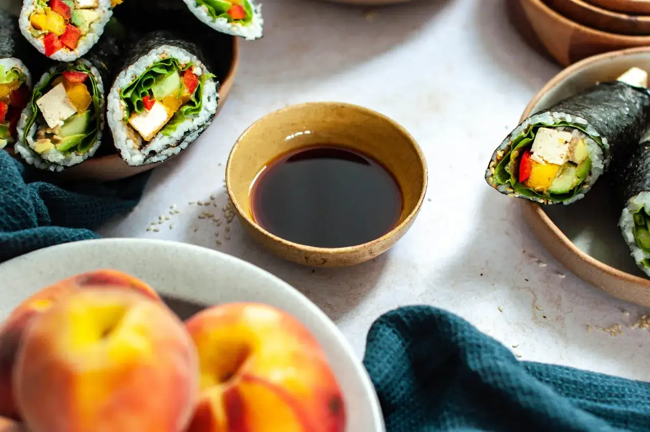 Z czym zrobić sushi wegetariańskie? Roladki z ryżem, nori, tofu, awokado, og&oacute;rkiem, papryką i sałatą. Obok miseczka sosu sojowego i brzoskwinie.