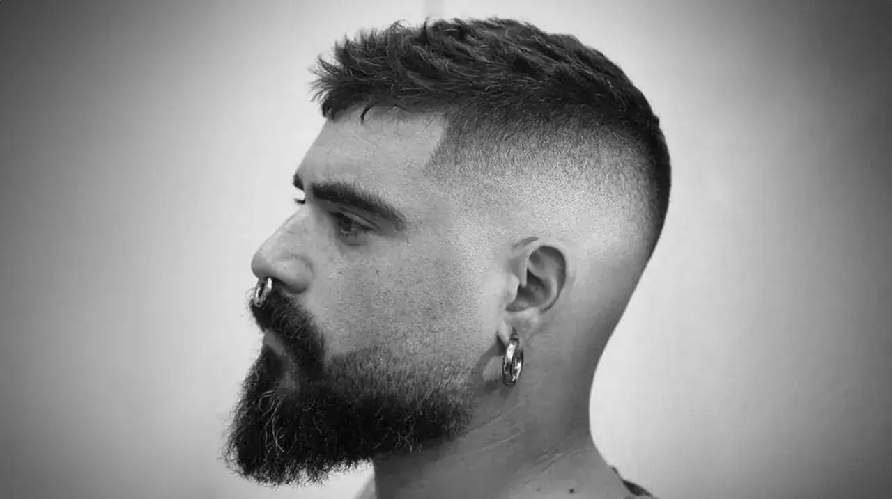 Mężczyzna z brodą i kolczykiem w nosie, z modnym, dłuższym buzz cut i cieniowanymi bokami.