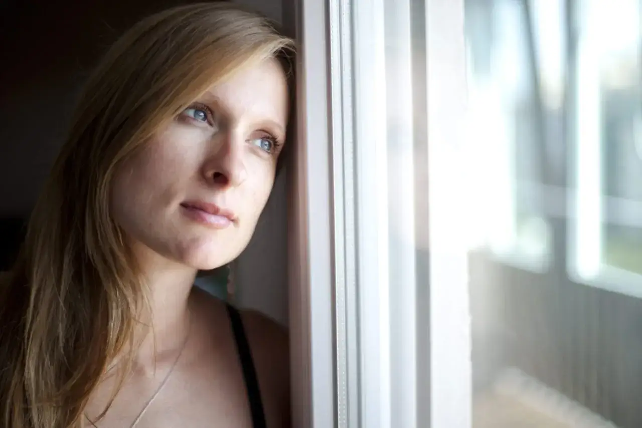 Blondynka opiera się o okno, wpatrując się w dal. W jej oczach widać tęsknotę za kimś.
