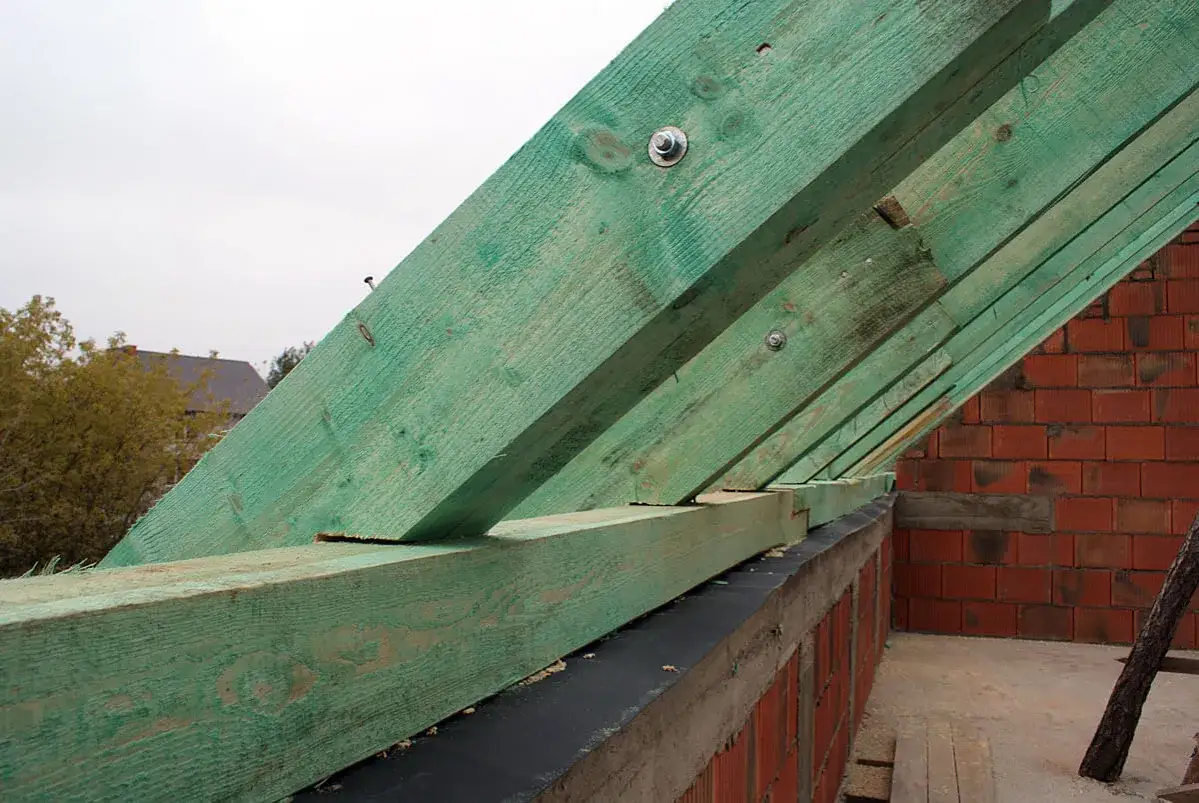 Solidne mocowanie krokwi do murłaty, zielone drewno konstrukcyjne na tle czerwonej cegły.