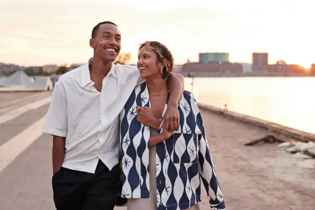 Uśmiechnięty mężczyzna obejmuje kobietę, spacerując nad wodą. Widać, jak facet zabiega o kobietę, okazując jej czułość i radość.