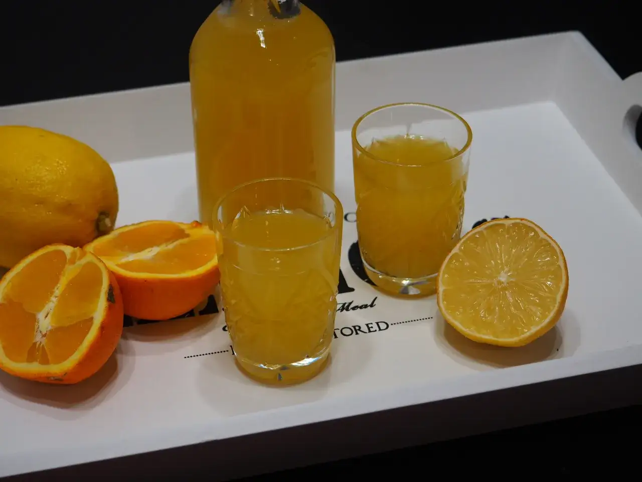 Jak przygotować domową wódkę smakową (cytrynówka) w 24 godziny? Na tacy butelka i szklanki z żółtym płynem, cytryny i pomarańcze.