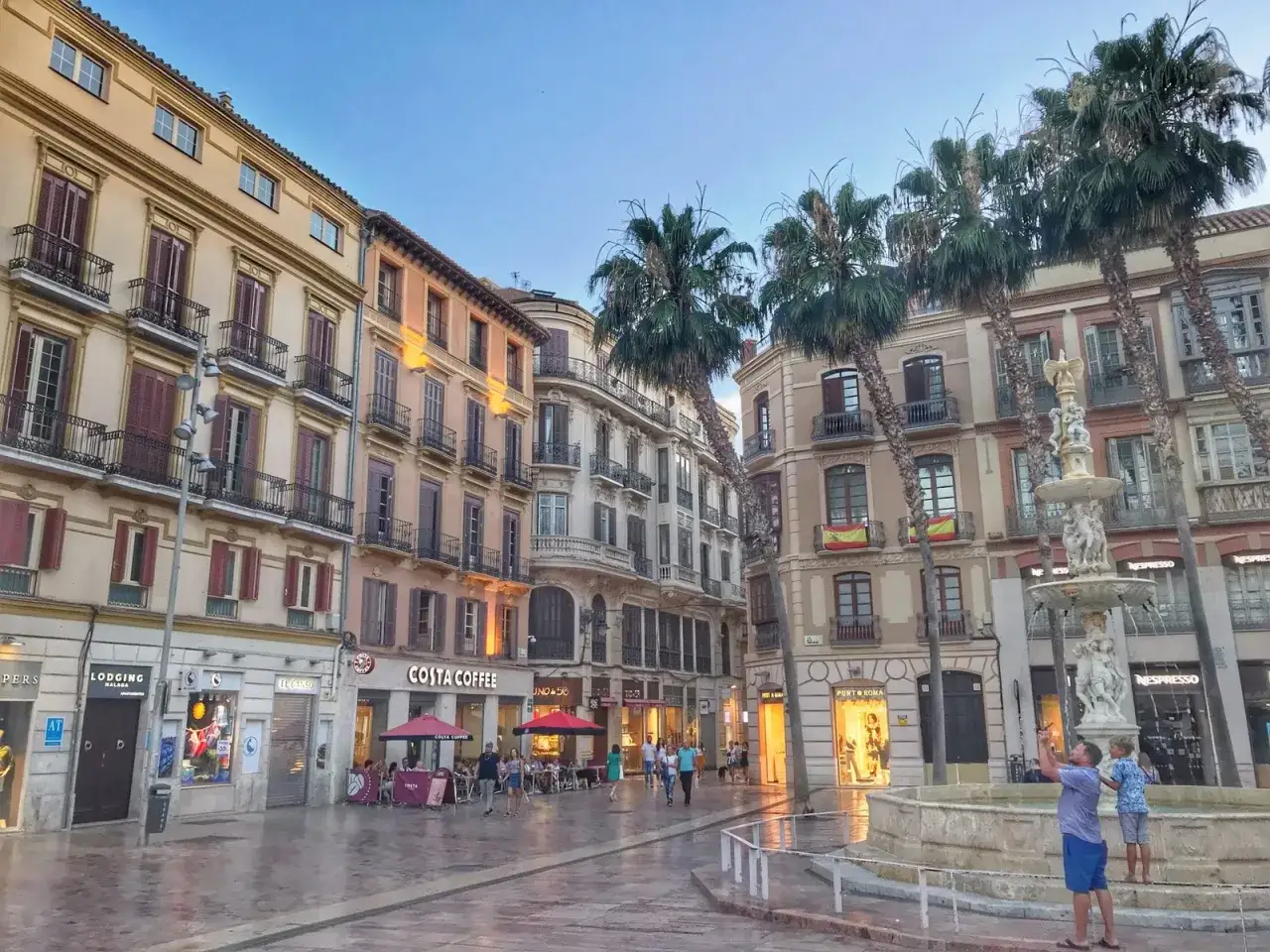 Malaga jest bezpieczna, o czym świadczy spokojny wieczór na placu z fontanną, palmami i ludźmi spacerującymi przy kawiarniach.