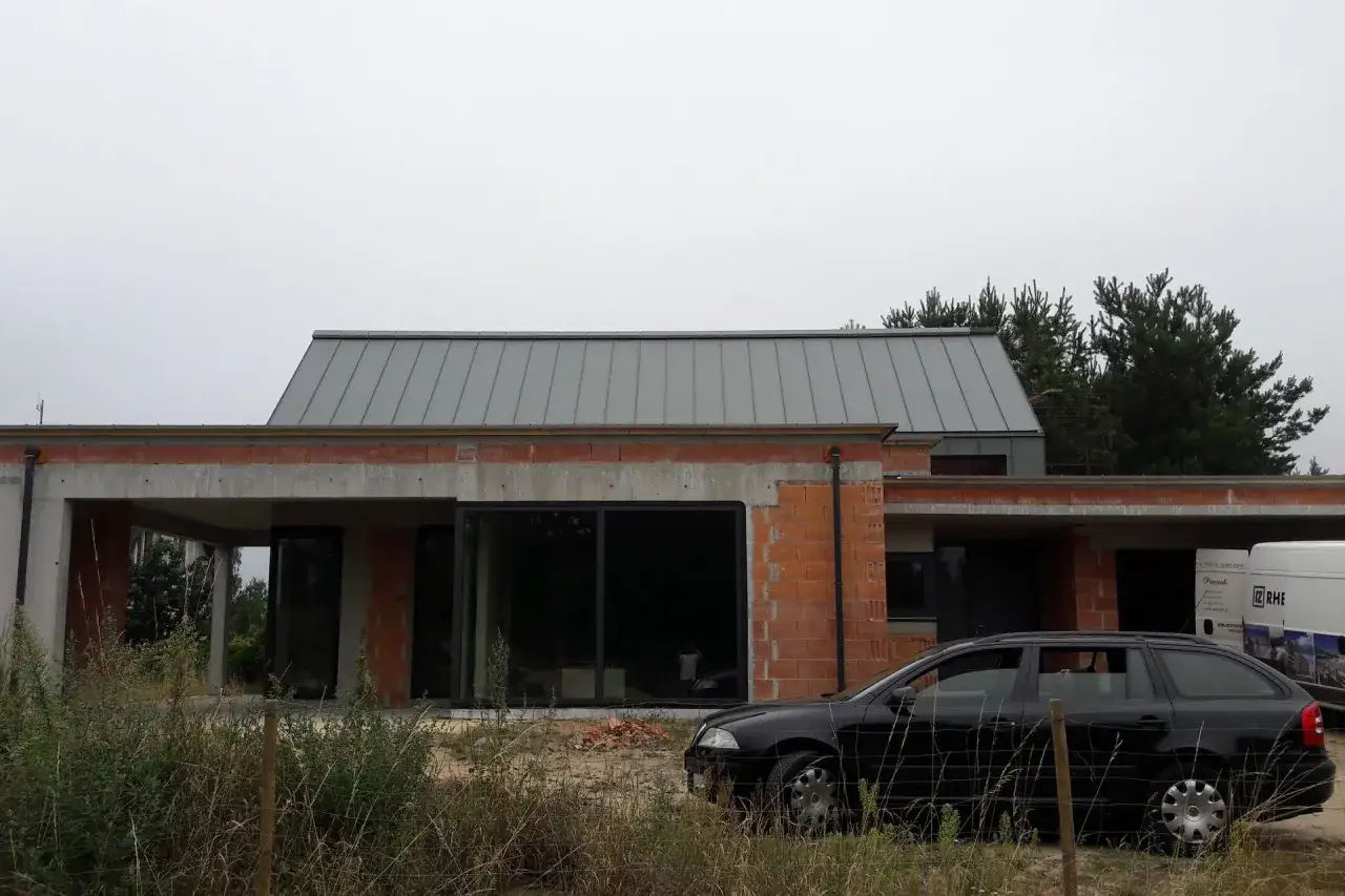 Nowoczesny dom w budowie z szarym dachem i ukrytą rynną. Obok czarny samoch&oacute;d i biała furgonetka.