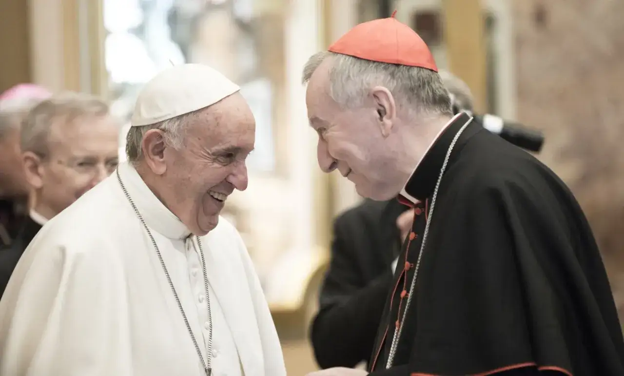 Papież Franciszek śmieje się, rozmawiając z kardynałem. Kto jest nad biskupem? To kardynał w czerwonej czapce i czarnej sutannie.