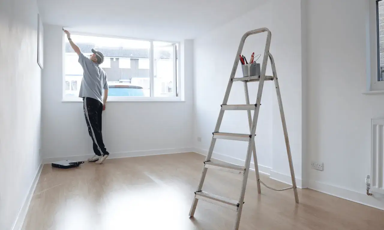 Mężczyzna maluje sufit w pokoju w stanie deweloperskim. Na drabinie stoją pędzle, a na podłodze kuweta z farbą.