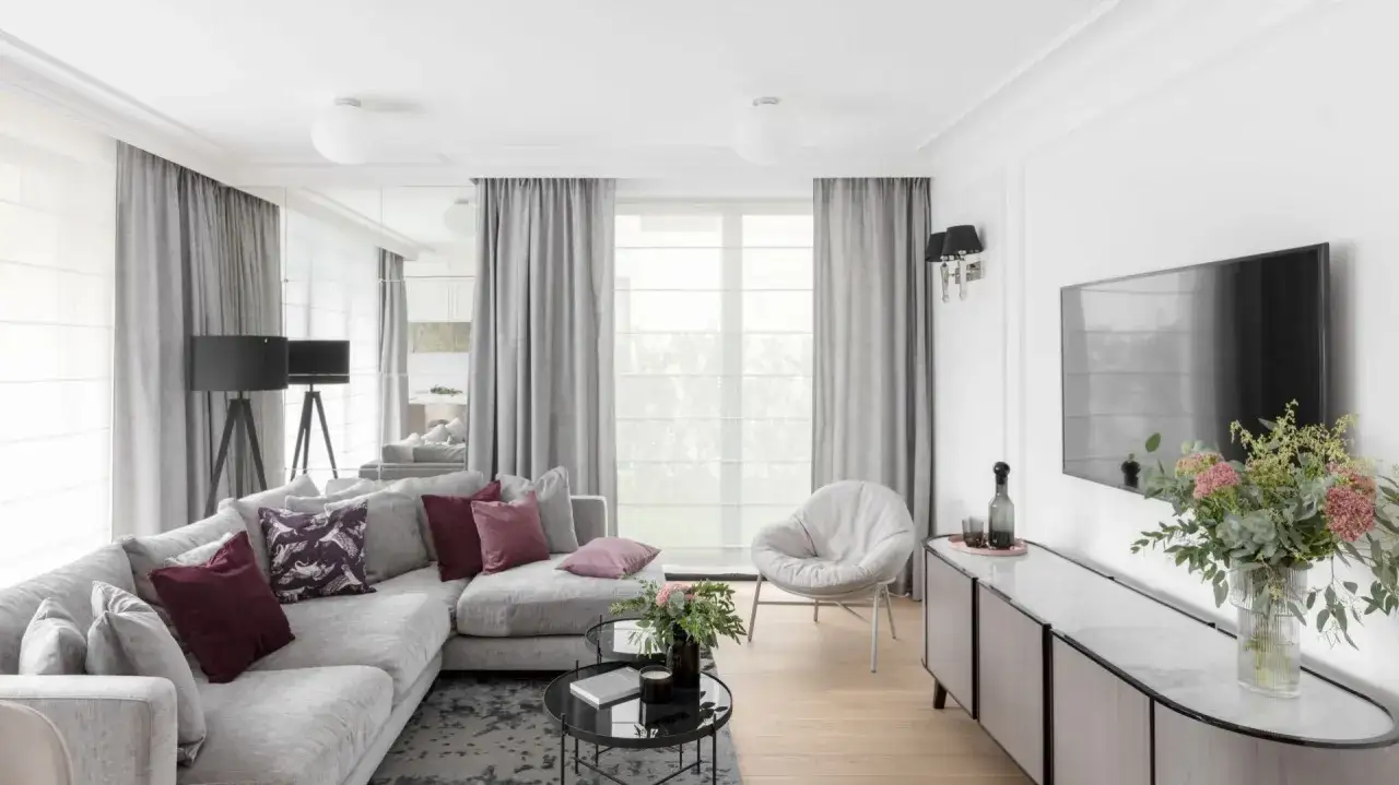 Elegancki salon z szarymi firanami, wygodną kanapą i okrągłym fotelem. Lustra odbijają światło, tworząc przestronną atmosferę.