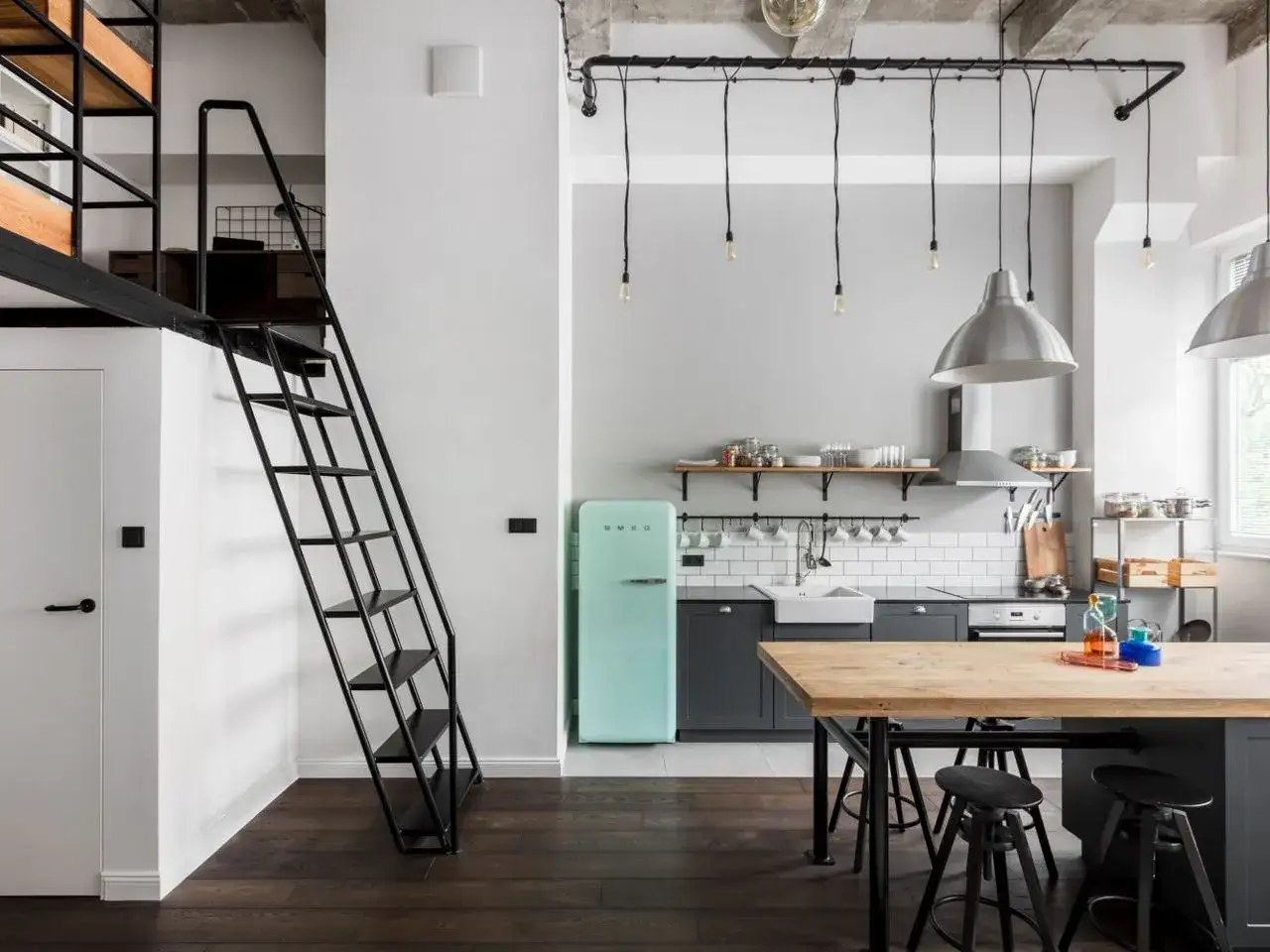 Industrialna kuchnia z miętową lod&oacute;wką i czarne kable na suficie tworzą loftowy klimat. Drewniany st&oacute;ł i metalowe schody dodają charakteru.