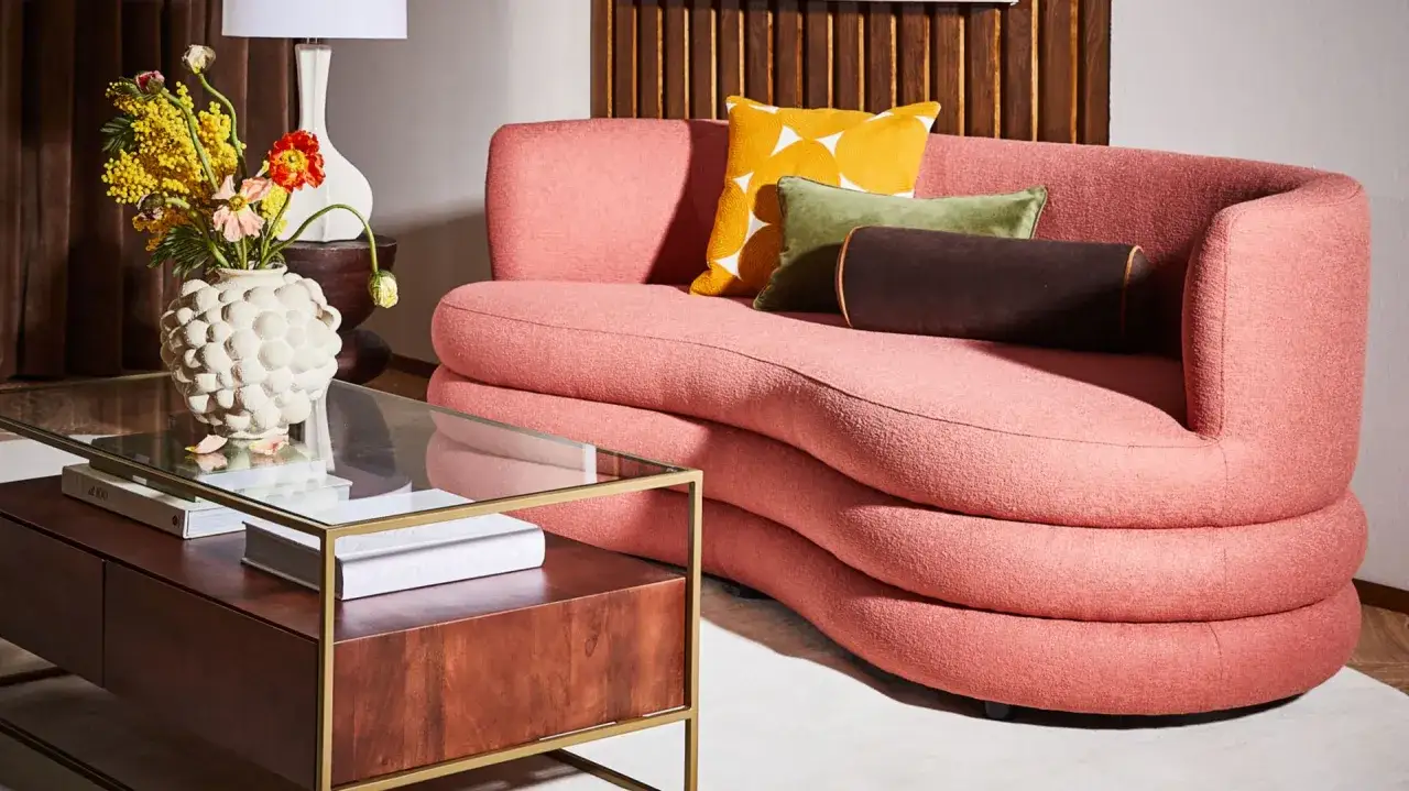 Przytulny **pok&oacute;j retro** z r&oacute;żową kanapą o falistych kształtach, szklanym stolikiem kawowym z książkami i wazonem z kwiatami.