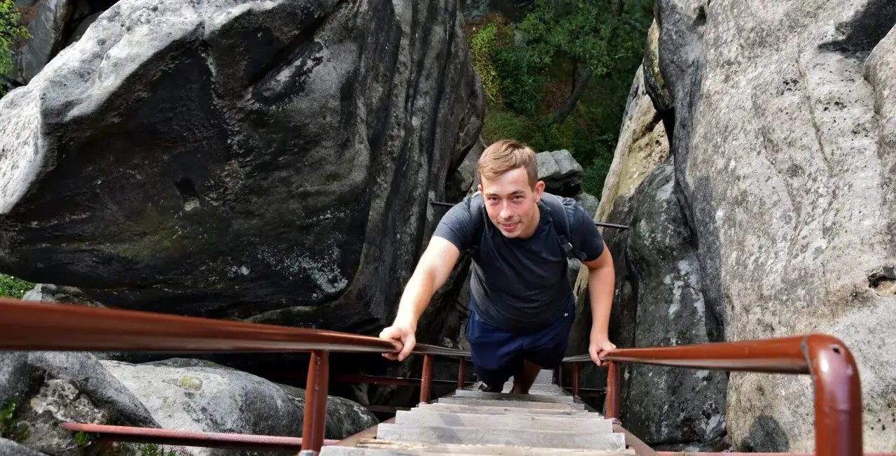 Młody mężczyzna wspina się po drewnianych schodach w skalnym mieście, chwytając się poręczy. To przygoda, kt&oacute;rą warto przeżyć!