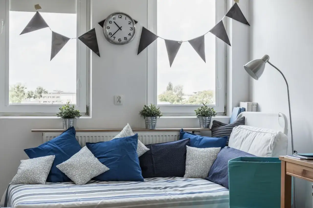 Urocze okno w pokoju dziecka aranżacje: ł&oacute;żko z niebieskimi poduszkami, girlanda z chorągiewek i zegar.