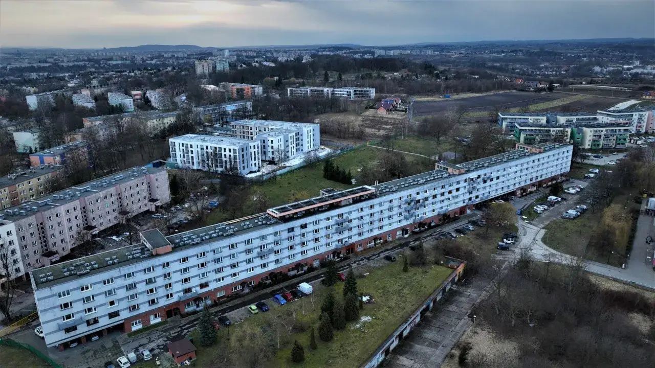 Długie bloki mieszkalne rozciągają się na wzgórzach, otoczone drzewami i parkingami. W tle widać inne budynki i zalesione tereny.
