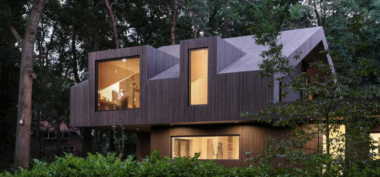 Nowoczesny dom w lesie z drewnianą elewacją i dużymi oknami, w tym jedno przypominające kształtem okno bawole oko, przez które widać ciepłe światło.