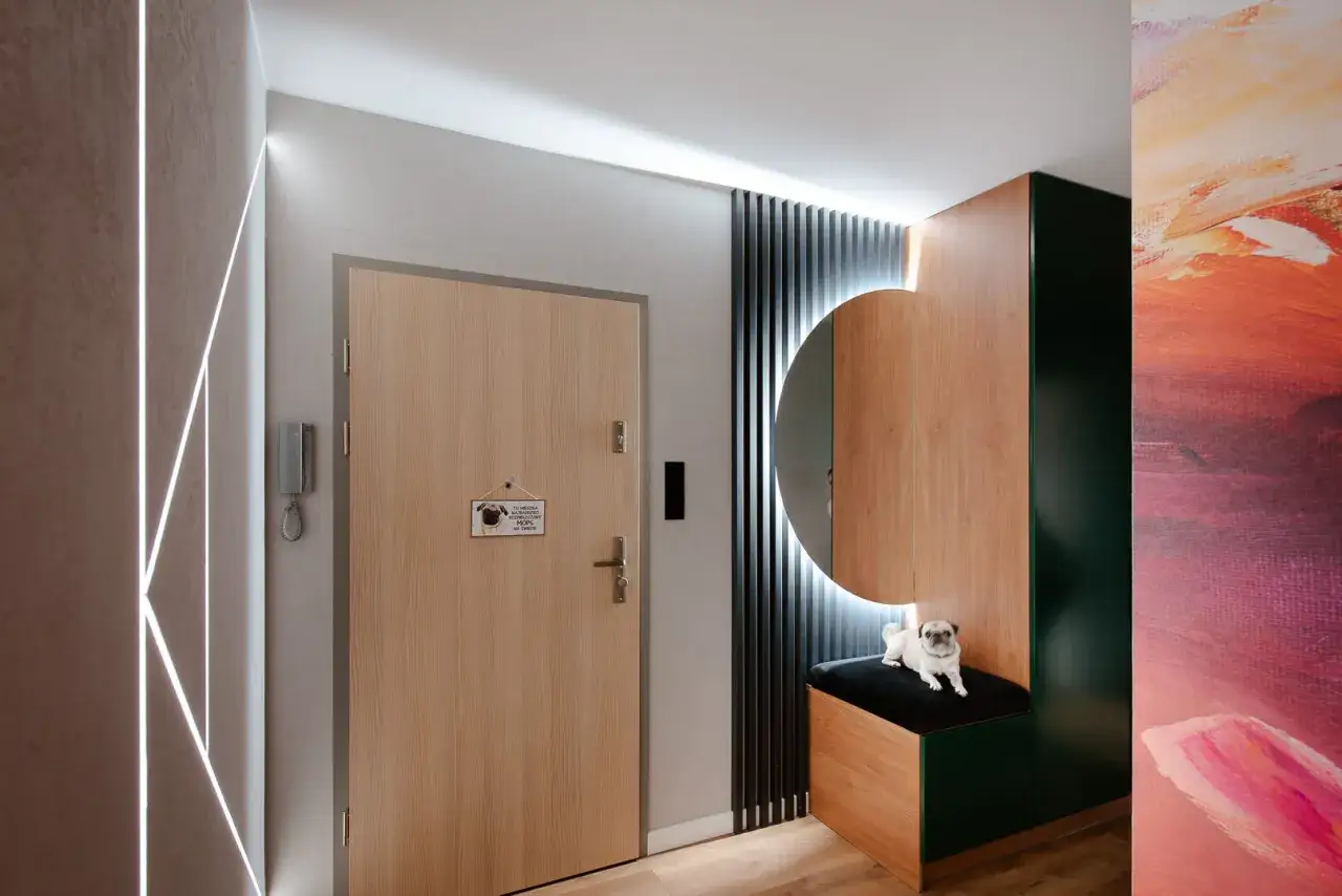 Nowoczesne lamele w przedpokoju tworzą stylową aranżację z drewnianymi frontami i okrągłym lustrem. Uroczy mops dodaje przytulności.