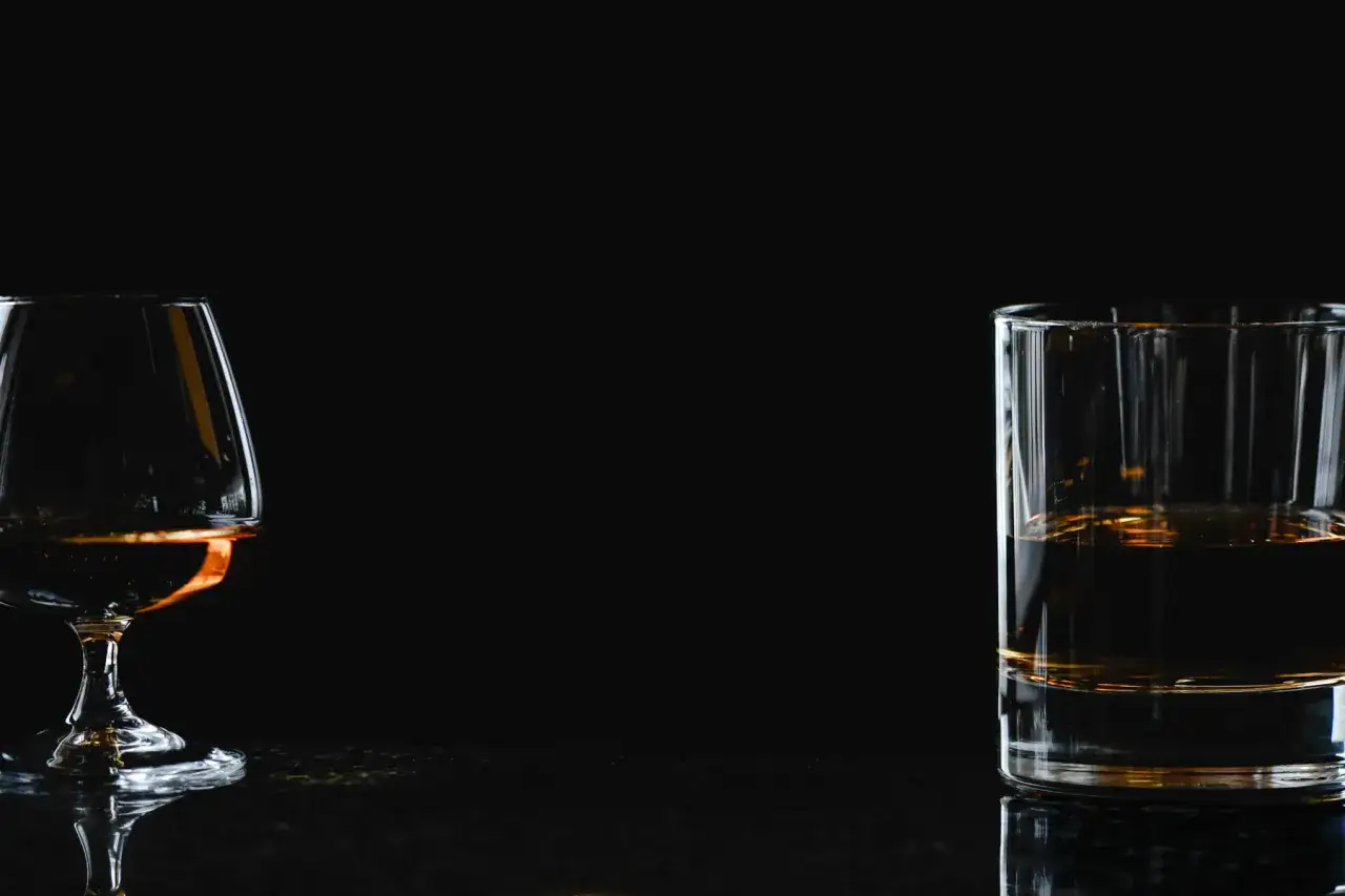 Kieliszek z brandy i szklanka z whisky stoją na ciemnym blacie, odbijając się w jego gładkiej powierzchni.